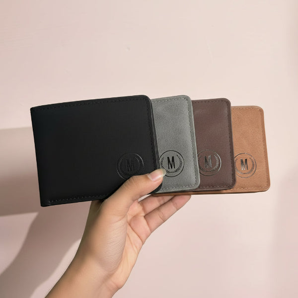A hand holds four slim bifold wallets in different colors: black, gray, brown, and tan. Each wallet features an embossed circular monogram logo with the letter 'M'.
