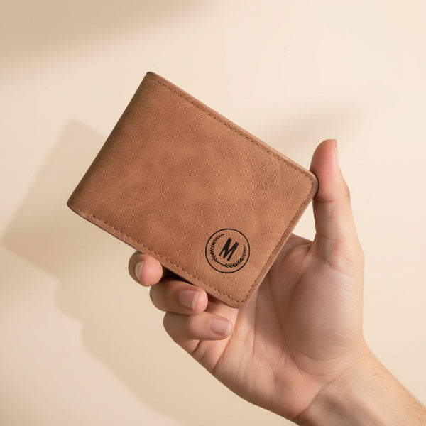 A hand holds four slim bifold wallets in different colors: black, gray, brown, and tan. Each wallet features an embossed circular monogram logo with the letter 'M'.