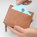 A tan leather zip-around card holder wallet being held by two hands, with a light blue card partially inserted into the top card slot.