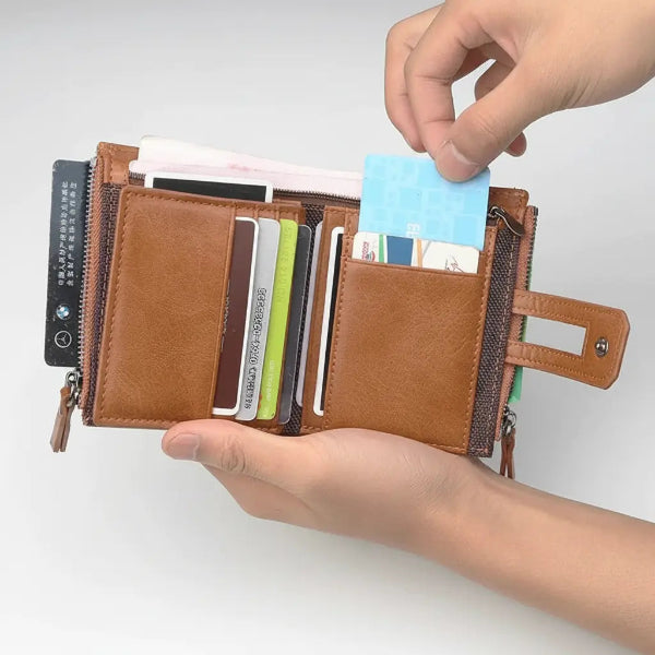A brown faux-leather bi-fold wallet shown open, held by two hands against a light gray background. The wallet contains multiple card slots, a zippered compartment, and side pockets, with a light blue credit card being inserted into one of the slots.