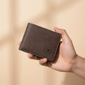 A person holds a brown faux-leather bifold wallet in front of a maroon leather sofa, with three other wallet color options (black, tan, and gray) lined up in the background. The front wallet features an embossed circular logo with the letter 'M'.