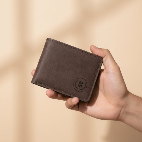 A person holds a brown faux-leather bifold wallet in front of a maroon leather sofa, with three other wallet color options (black, tan, and gray) lined up in the background. The front wallet features an embossed circular logo with the letter 'M'.
