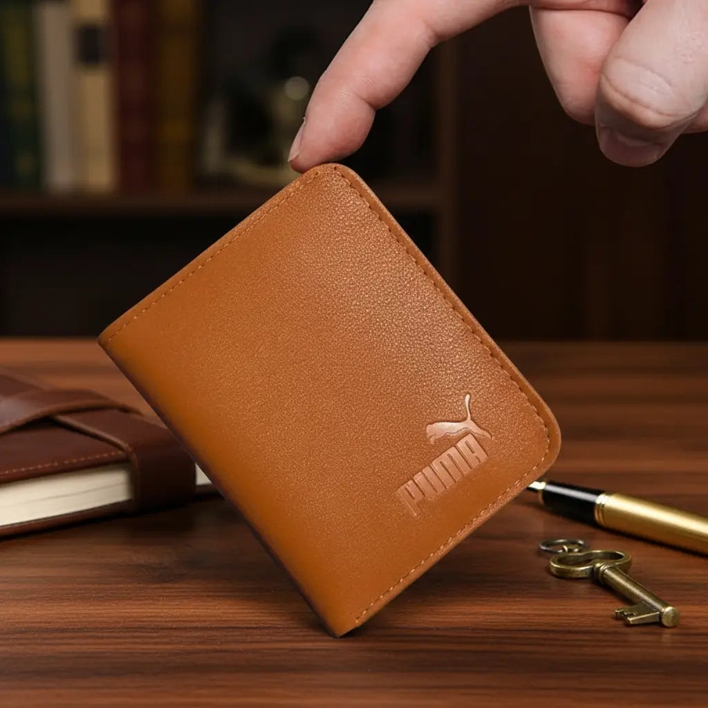 A tan Puma leather wallet held by a person's fingers on a wooden desk, with a pen, keys, and a notebook nearby.