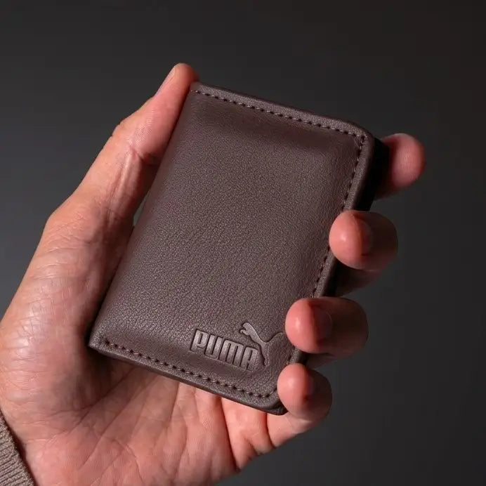 A hand holding a small brown leather wallet with the Puma logo embossed on the front, set against a dark gray background.